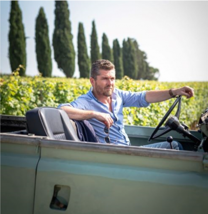 winemaker Nicolas Audebert drives an open-top 4-wheel drive along the edge of Berliquet's vineyards. Cyprus trees stand in the background