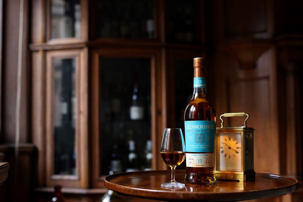 A bottle of Glenmorangie 13-year-old Cognac Finish stands next to a large dram in a stemmed glass. The scene is in No.3, with wooden pannels and a small clock.
