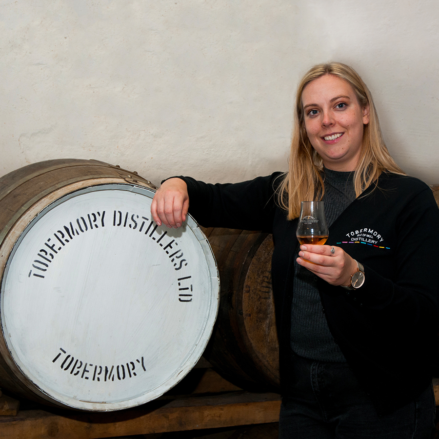 A photo of Cara Gilbert at Tobermory Distillery. She is holding a glass of whisky, with one arm propped on a barrel imprinted with the distillery name. 