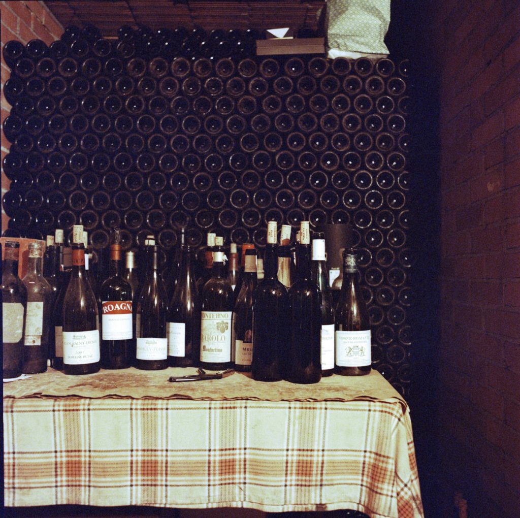 A photograph showing dozens of Barolo bottles sitting atop a checked table, against the backdrop of a producer's cellar in Piedmont. 