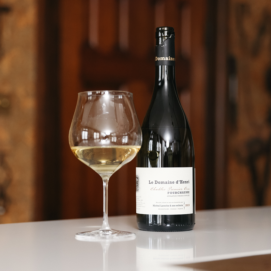 A bottle of Le Domaine d'Henri's Chablis sits atop a white table next to a Burgundy glass holding a little of the golden-coloured wine, against a wood backdrop