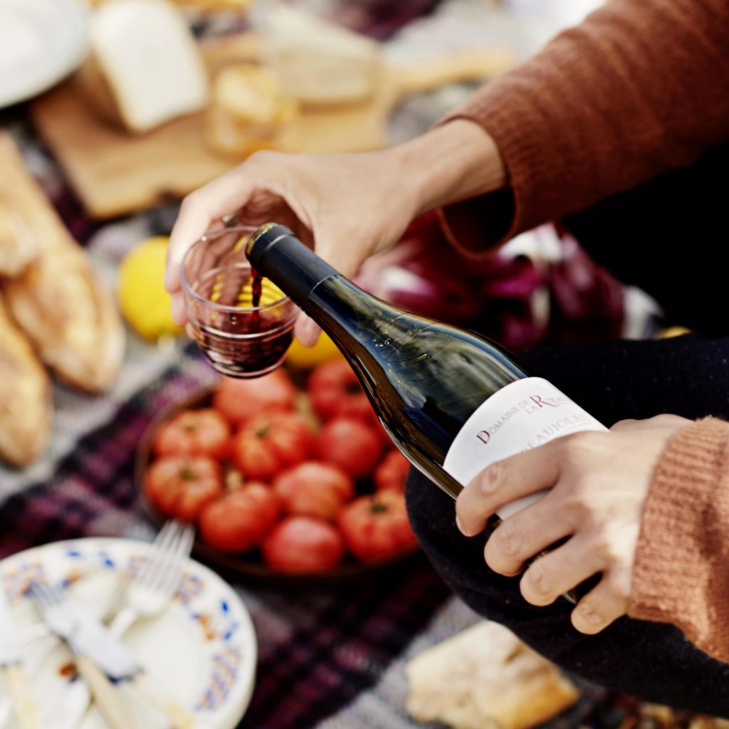 Someone pouring red wine into a glass, with picnic food in the background.
