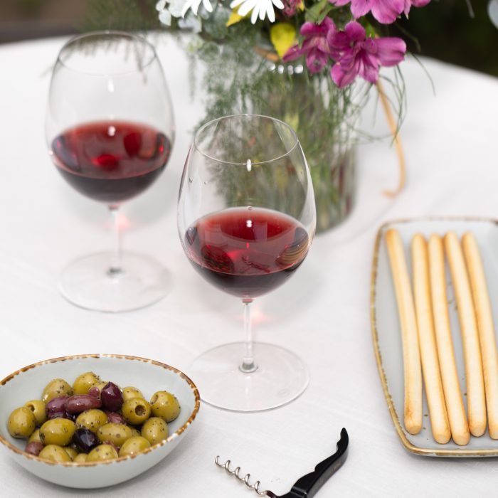 Two glasses of red wine on a table with some olives and breadsticks.